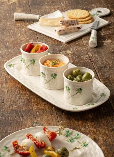 A rustic cheese and appetizer spread featuring various cheeses, olives, sun-dried tomatoes, and dips served in white ceramic dishes tapas dishes with a green shamrock on a wooden table.